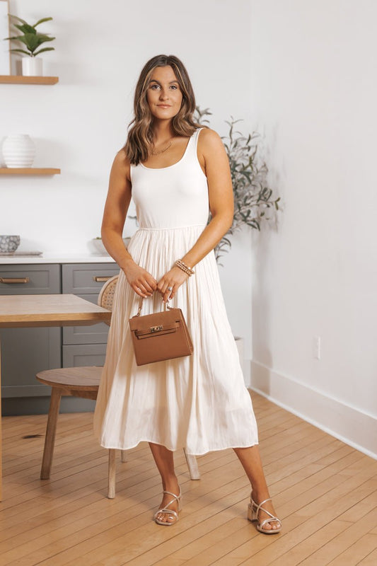 Woman wears a Scoop Neck Pleated Skirt Midi Tank Dress, holding a brown handbag in a minimal room with light wood floors.
