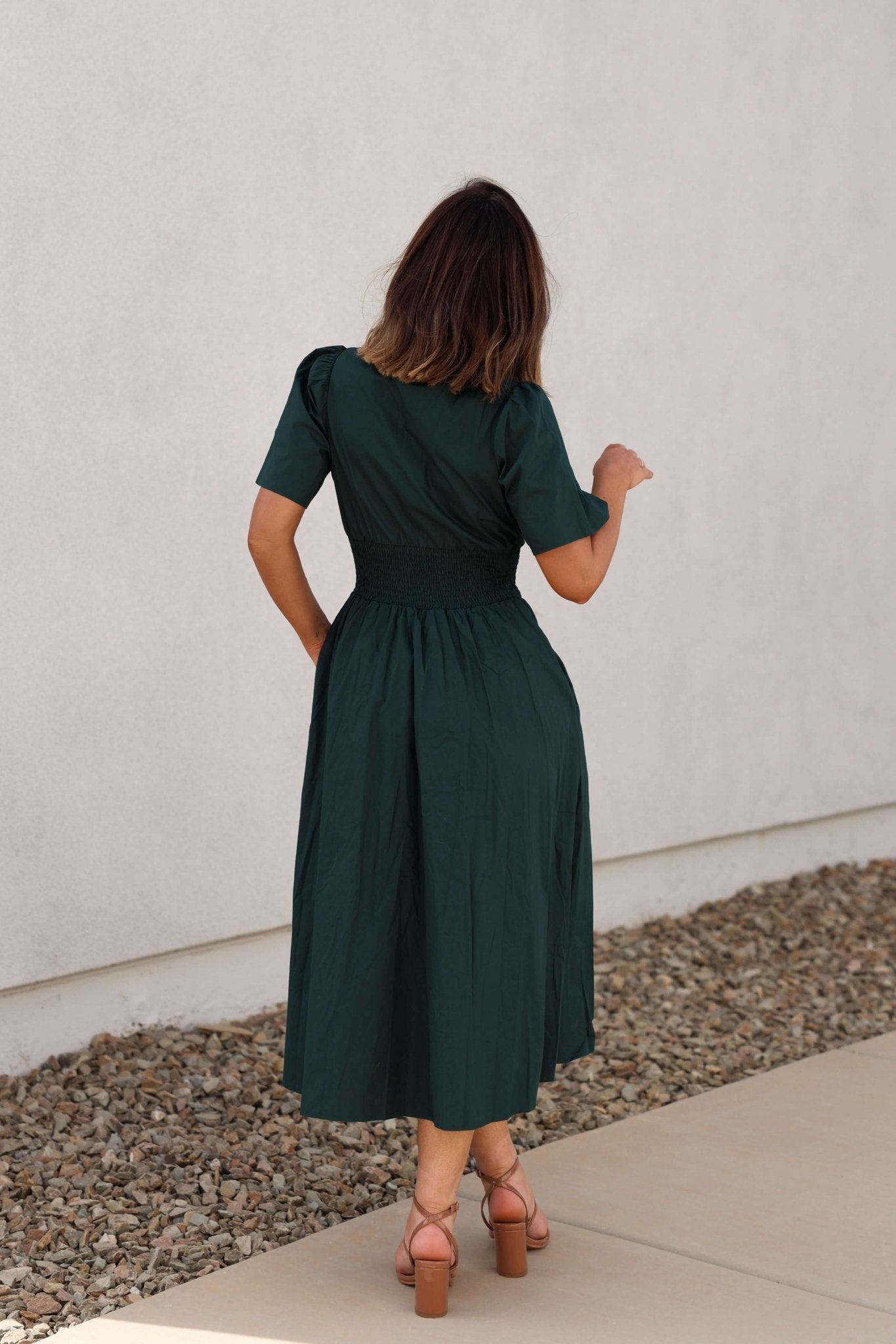 A woman wears the Sea Green Smocked Zip Midi Dress - FINAL SALE with tan heels, standing outside on a sidewalk, facing away.