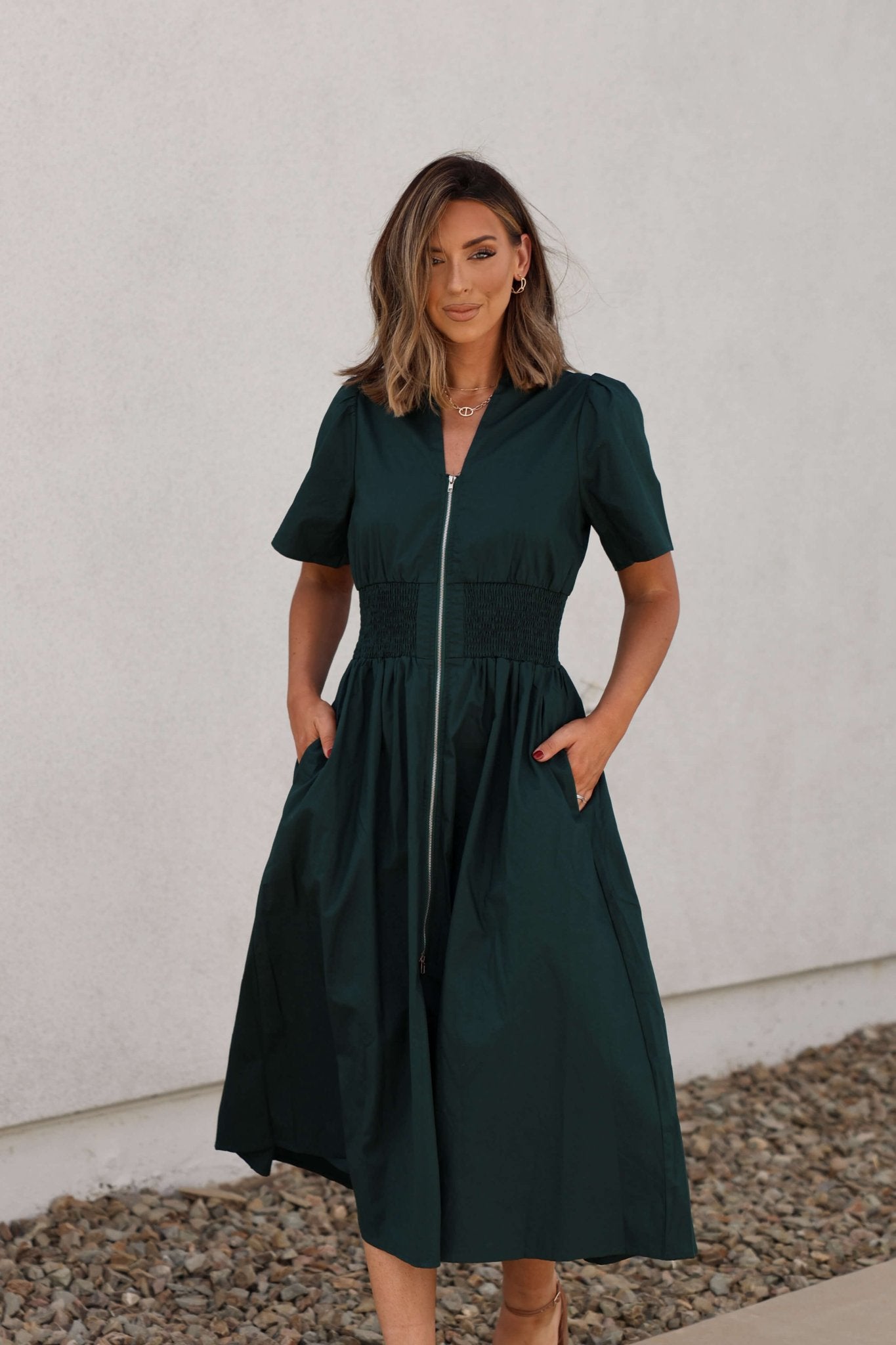 Woman wearing the Sea Green Smocked Zip Midi Dress stands outdoors with hands in pockets, posing on a stone path.