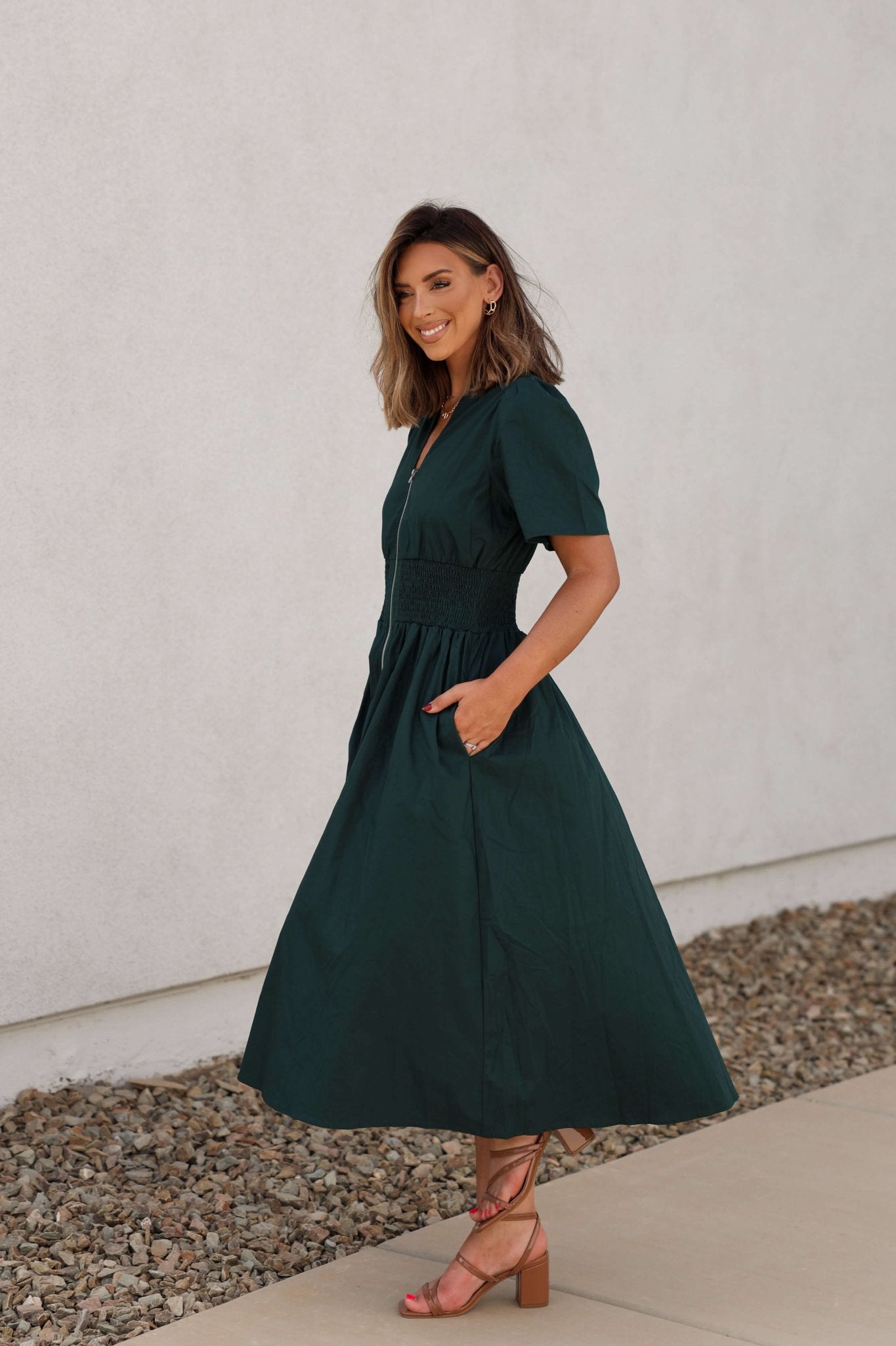 Woman in the Sea Green Smocked Zip Midi Dress, smiling by a stone border on the sidewalk.