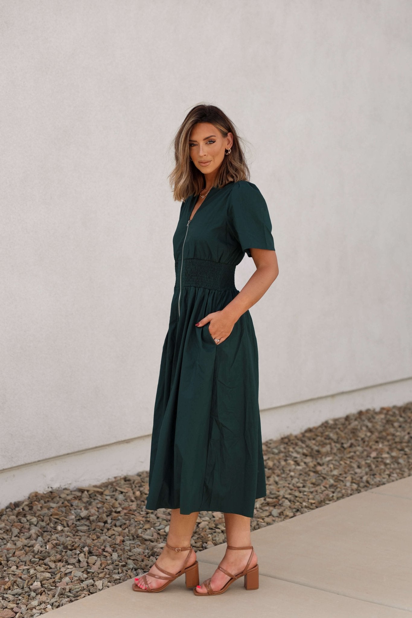 Woman in a Sea Green Smocked Zip Midi Dress - FINAL SALE, standing outdoors on a sidewalk by a light wall.