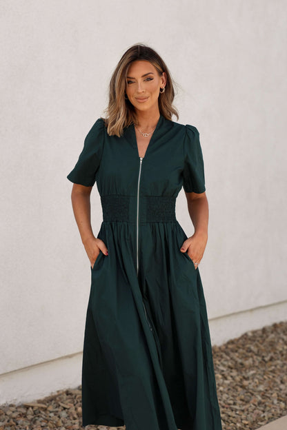 Woman in a Sea Green Smocked Zip Midi Dress - FINAL SALE stands on a pebble path by a white wall.