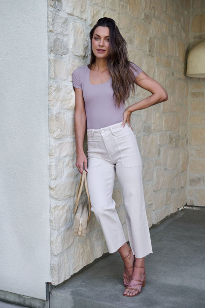 Woman with long wavy hair in a lavender top, Sea Salt Grey High Rise Wide Leg Jeans, and sandals holds a beige handbag by a stone wall.