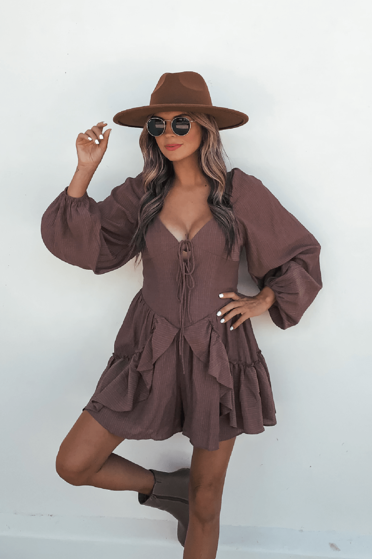 Woman models the Shay Mocha Ruffled Sweetheart Romper with long bubble sleeves, brown hat, sunglasses, and ankle boots.