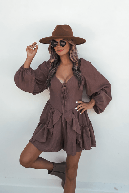 Woman in the Shay Mocha Ruffled Sweetheart Romper, brown hat, and round sunglasses poses against a plain white background.