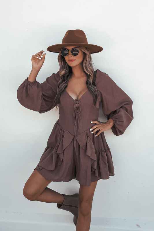 Woman in the Shay Mocha Ruffled Sweetheart Romper, brown hat, and round sunglasses poses against a plain white background.