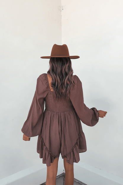 Woman with wavy hair wears the Shay Mocha Ruffled Sweetheart Romper, standing back to camera against a white wall.