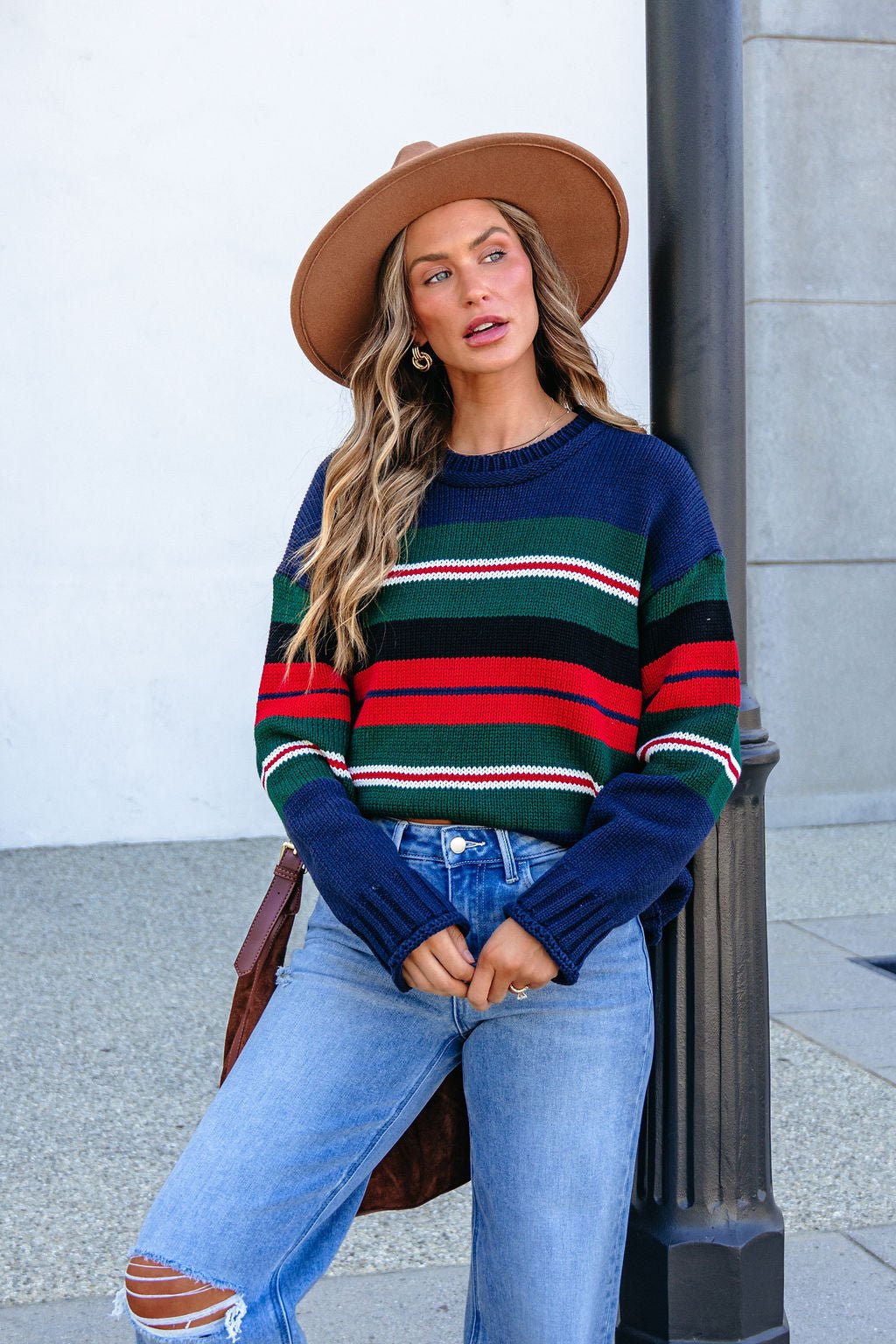 Woman wears the SHE AND SKY ST1207 navy striped sweater, ripped jeans, and brown hat by a lamp post with a light backdrop.
