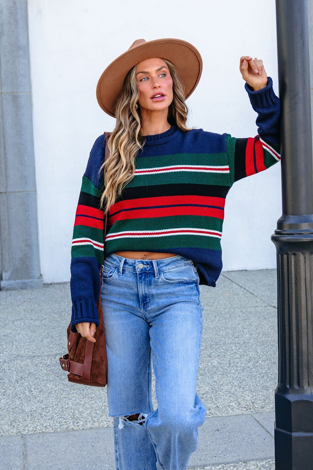 Woman in SHE AND SKY ST1207 navy striped sweater, ripped jeans, and hat stands by a lamppost with a brown bag, looking aside.