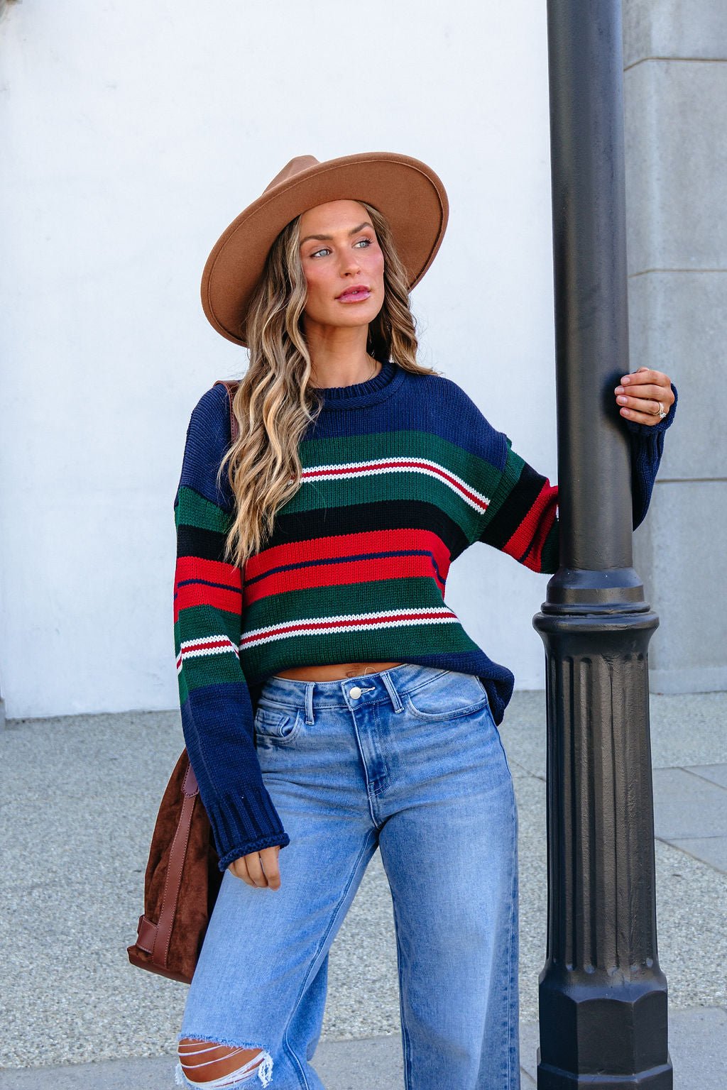 Woman in a SHE AND SKY ST1207 navy striped sweater, ripped jeans, and hat stands outdoors holding a black lamp post, looking aside.