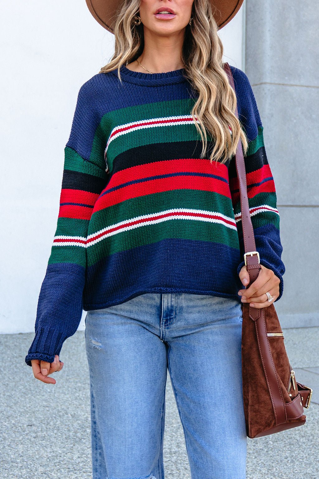 Woman in SHE AND SKY ST1207 navy striped sweater, jeans, brown bag, and hat stands outdoors with one hand slightly raised.
