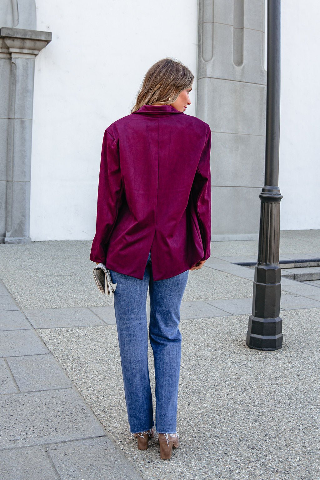 A woman wears a SHE AND SKY ST1741 wine suede oversized blazer jacket with blue jeans, standing on a sidewalk facing away.