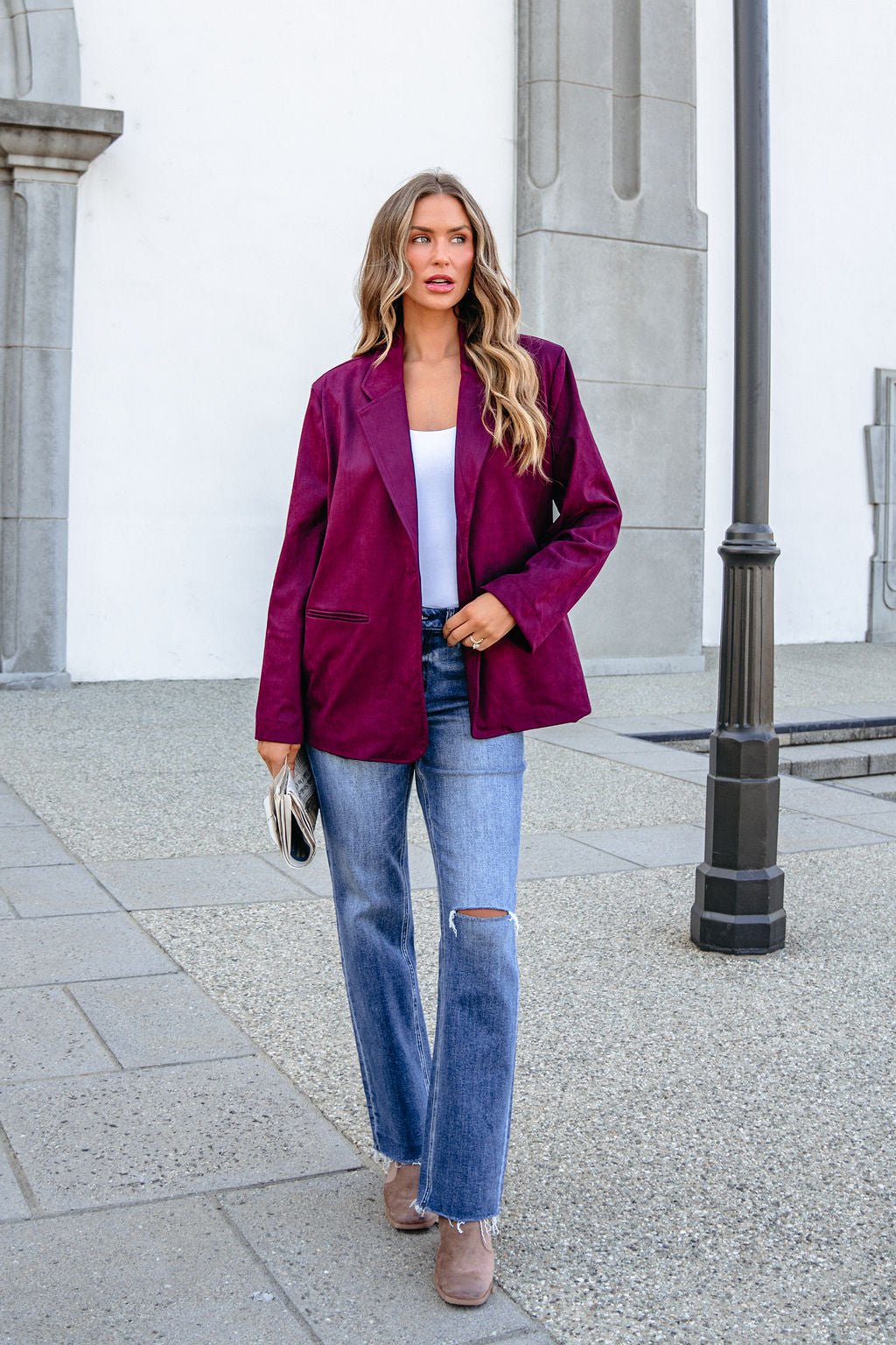 Woman in a SHE AND SKY ST1741 wine suede blazer, white top, and ripped jeans walks on a sidewalk near a gray building holding a clutch.
