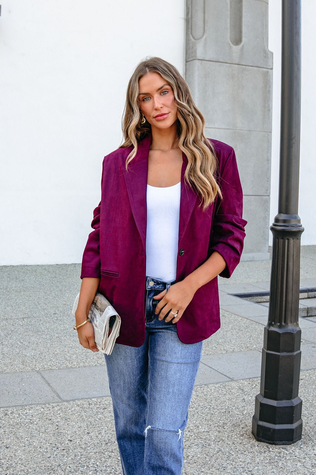 Woman in SHE AND SKY ST1741 wine suede oversized blazer, white top, and jeans holds a clutch near a lamp post by a white wall.