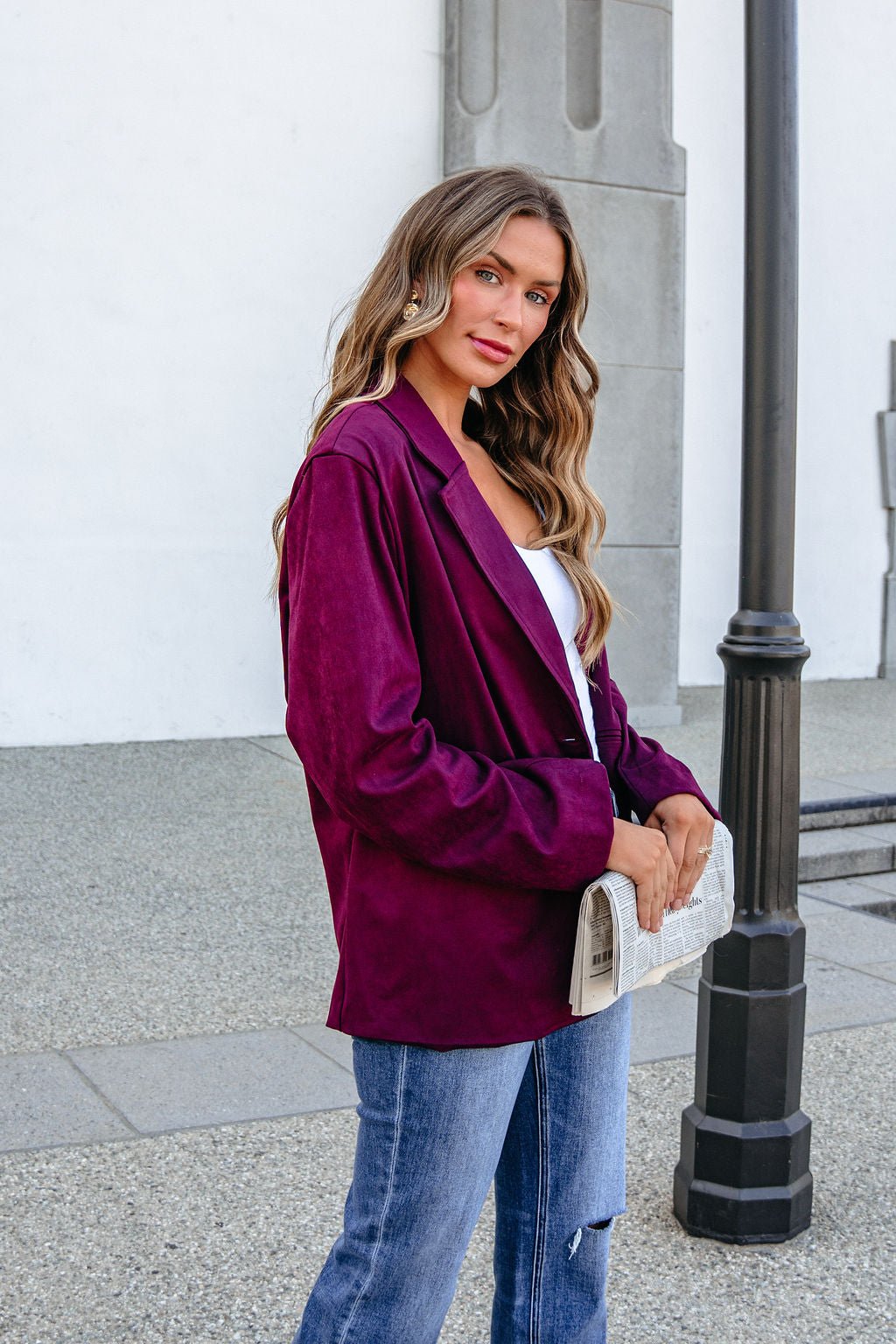 Woman in the SHE AND SKY ST1741 Wine Suede Oversized Blazer Jacket stands outdoors, holding a newspaper clutch by a lamp post.
