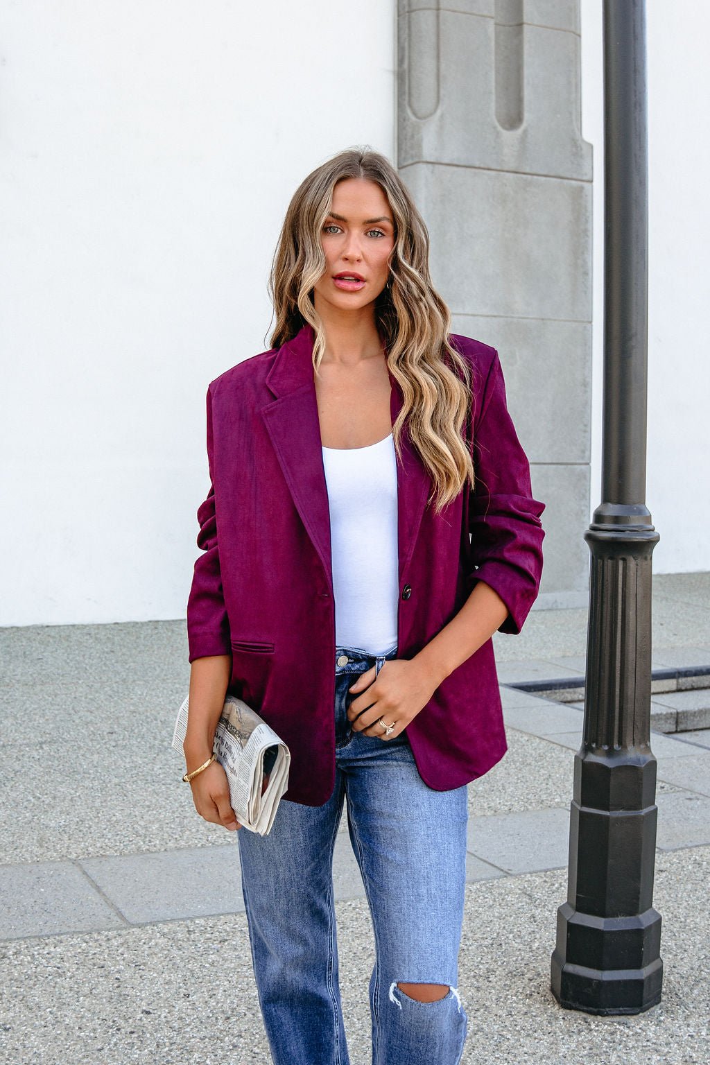 Woman in a wine suede oversized blazer jacket holds a beige clutch near a black streetlamp.