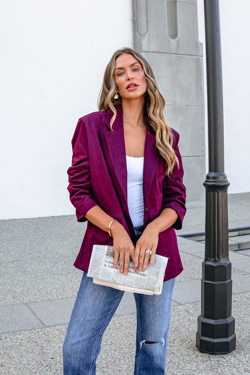 Woman with wavy hair in SHE AND SKY ST1741 Wine Suede Oversized Blazer Jacket holds a newspaper near a black lamppost outdoors.