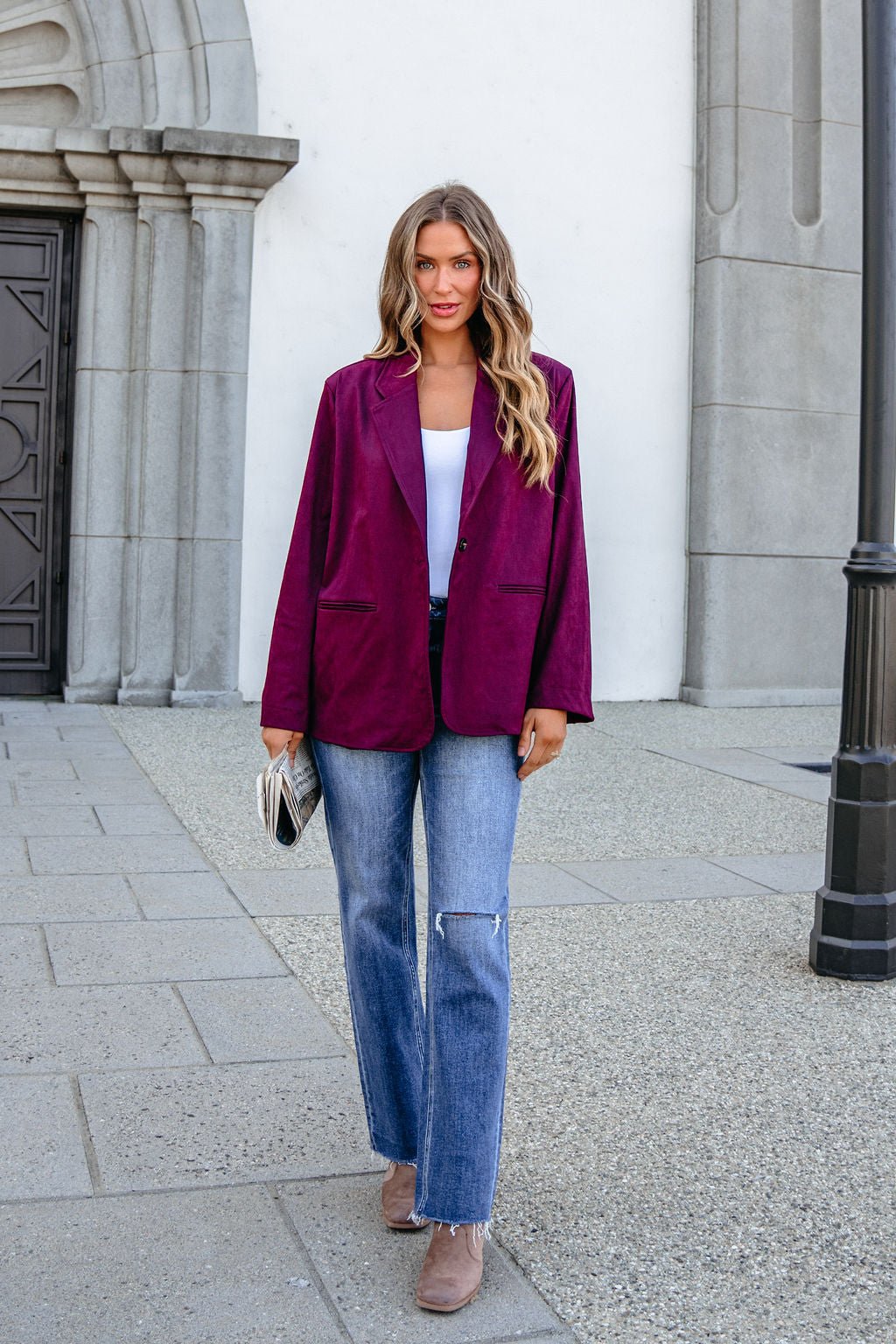 Woman in SHE AND SKY ST1741 wine suede oversized blazer, white top, ripped jeans, taupe boots outside by stone building with striped clutch.