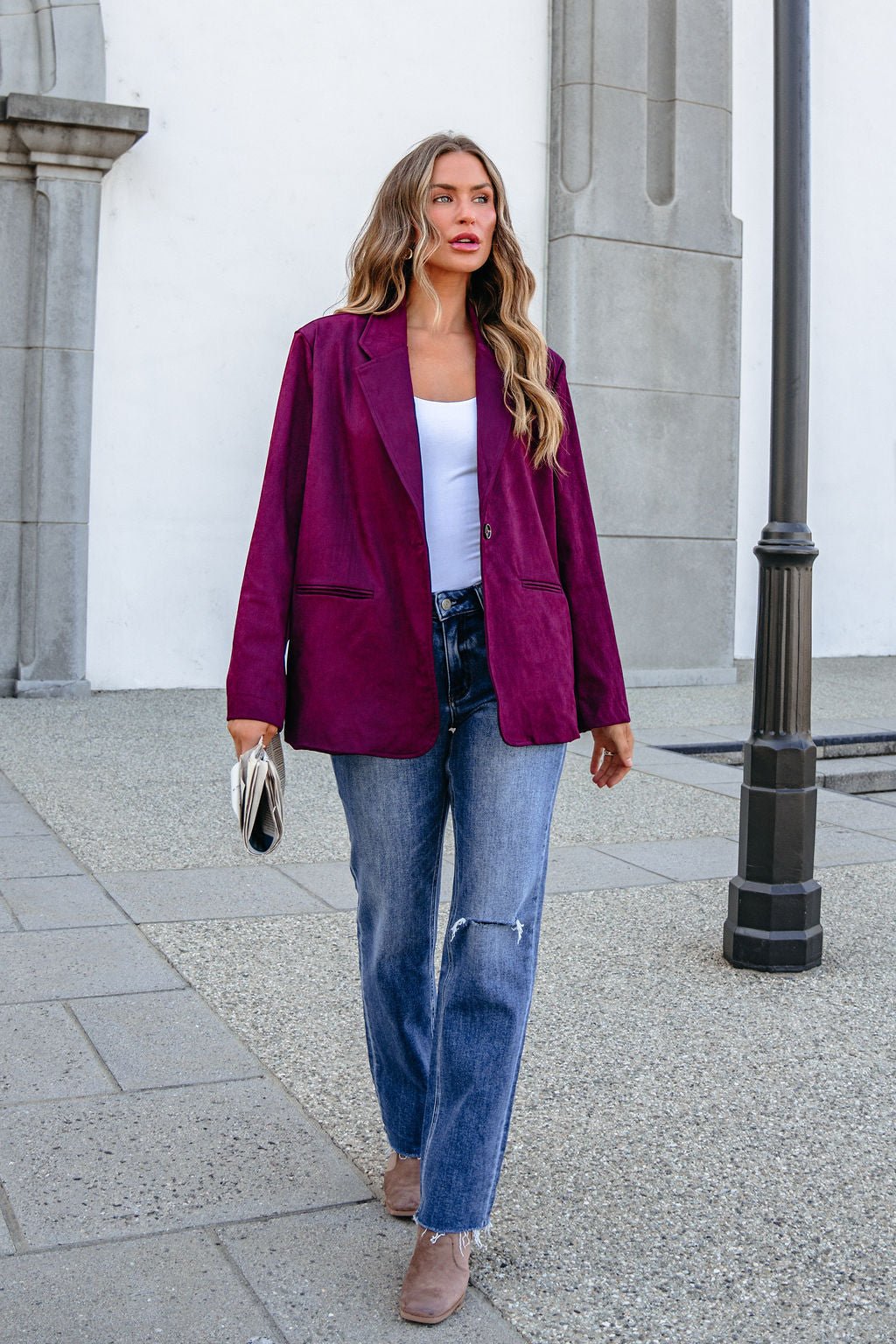 Woman in a SHE AND SKY ST1741 wine suede oversized blazer jacket, white top, and jeans walks outside holding a clutch.