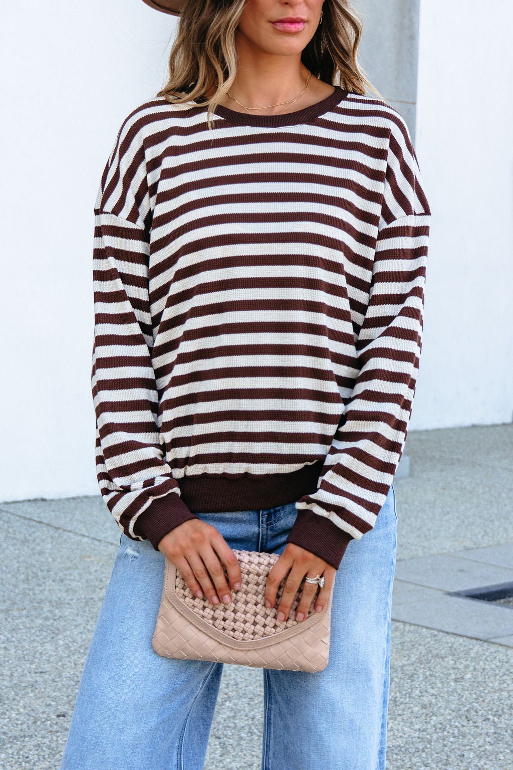 Woman in a SHE AND SKY ST1813 brown striped top, blue jeans, with a woven beige clutch, standing outdoors.