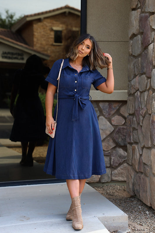 A woman wears a navy Collared Short Sleeve Midi Dress with a waist tie and tan ankle boots.