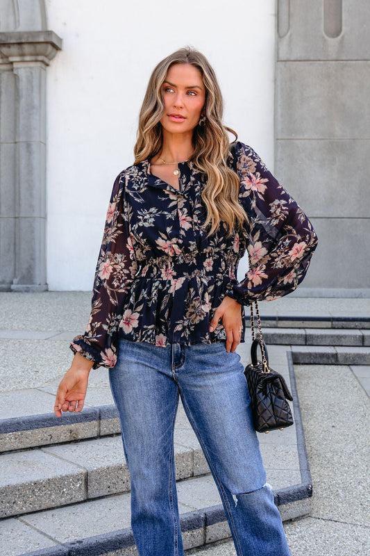 A woman wears a black Floral Long Sleeve Smock Top with sheer chiffon floral fabric, a smocked waist, and blue jeans.