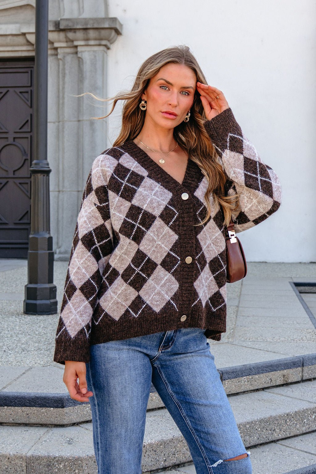 Woman in SHE AND SKY ST1999 brown argyle sweater and ripped jeans stands on steps, touching her hair and facing the camera.