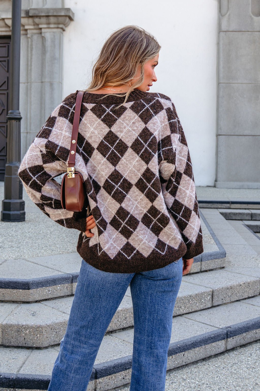 Woman with long hair wears the SHE AND SKY ST1999 brown argyle sweater and blue jeans, standing outdoors facing away.
