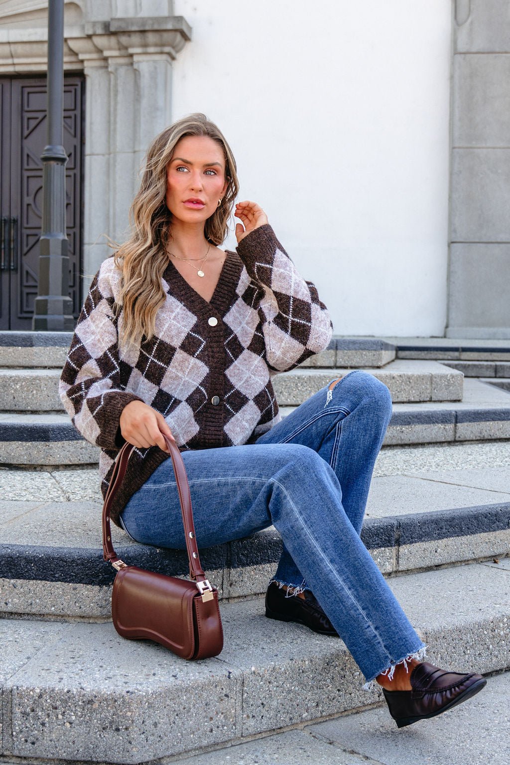 Woman on outdoor steps, wearing SHE AND SKY ST1999 brown argyle sweater, blue jeans, black shoes, holding a brown handbag.