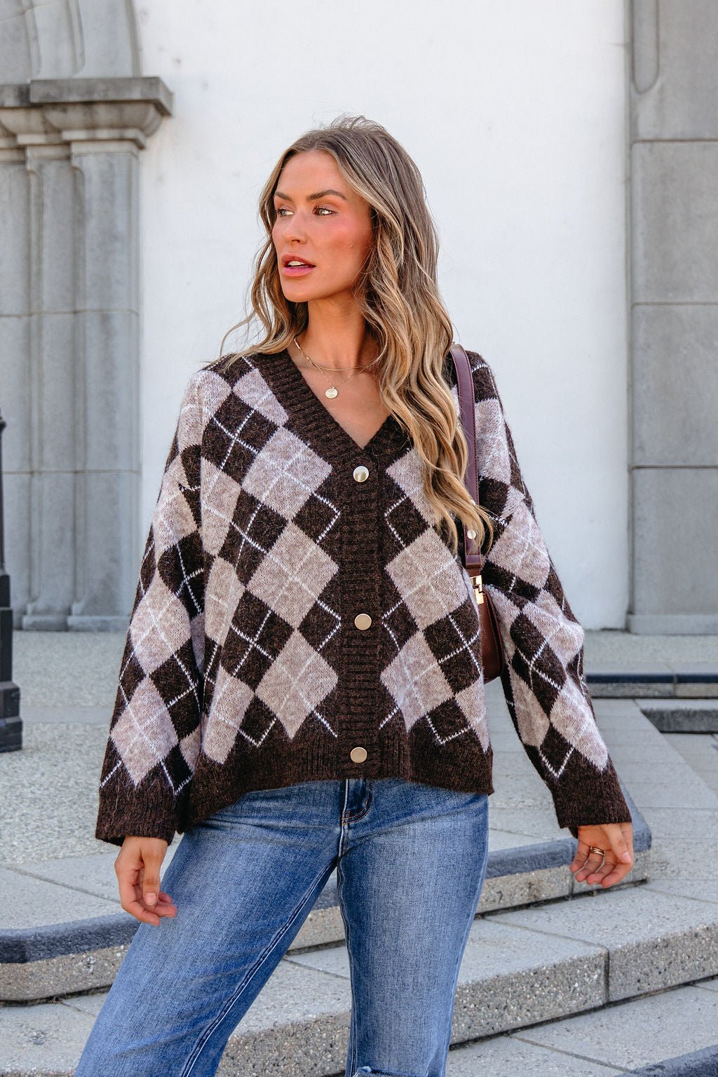 Woman wears the SHE AND SKY ST1999 brown argyle sweater and jeans, standing outdoors by a stone building, looking to the side.