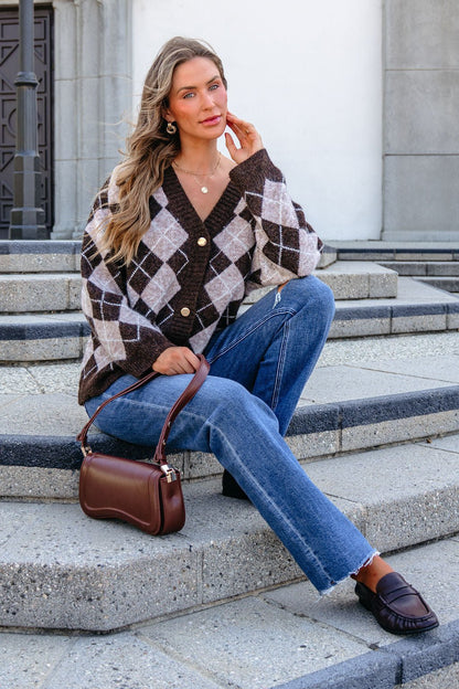 Woman on stone steps in SHE AND SKY ST1999 brown argyle sweater, blue jeans, loafers, holding a brown purse.