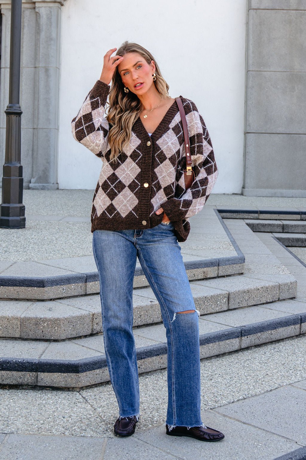 Woman in SHE AND SKY ST1999 brown argyle sweater, jeans, and loafers stands on stone steps with a brown shoulder bag.