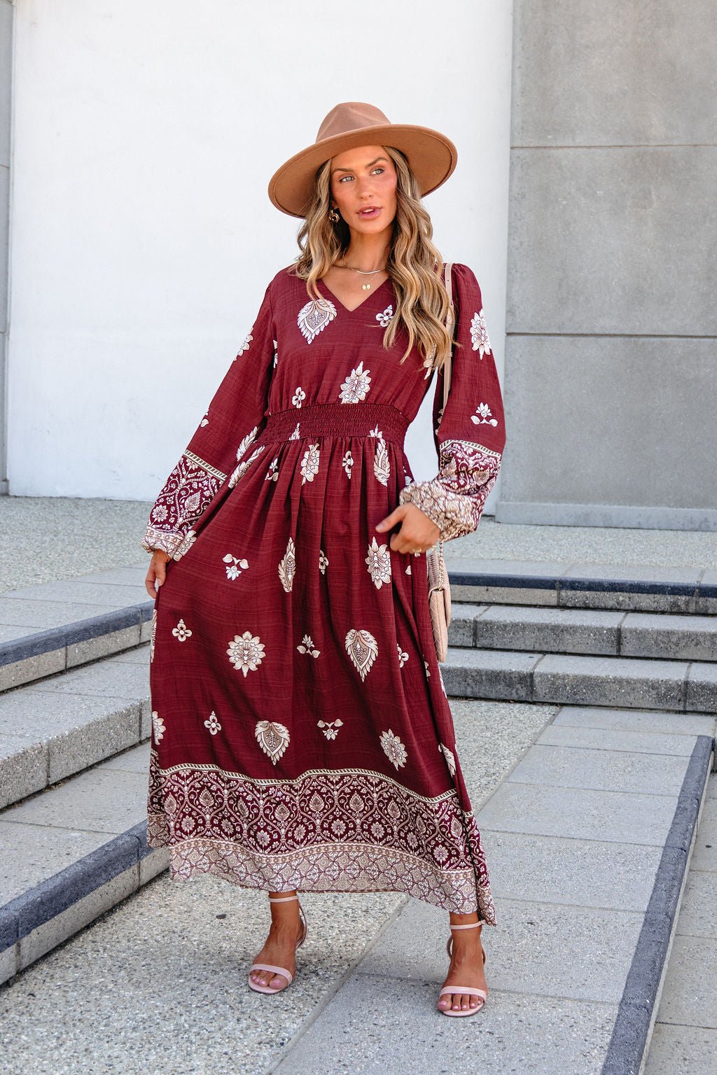 Woman in SHE AND SKY ST2144 brown printed maxi dress, tan hat, and sandals poses outdoors on concrete steps.