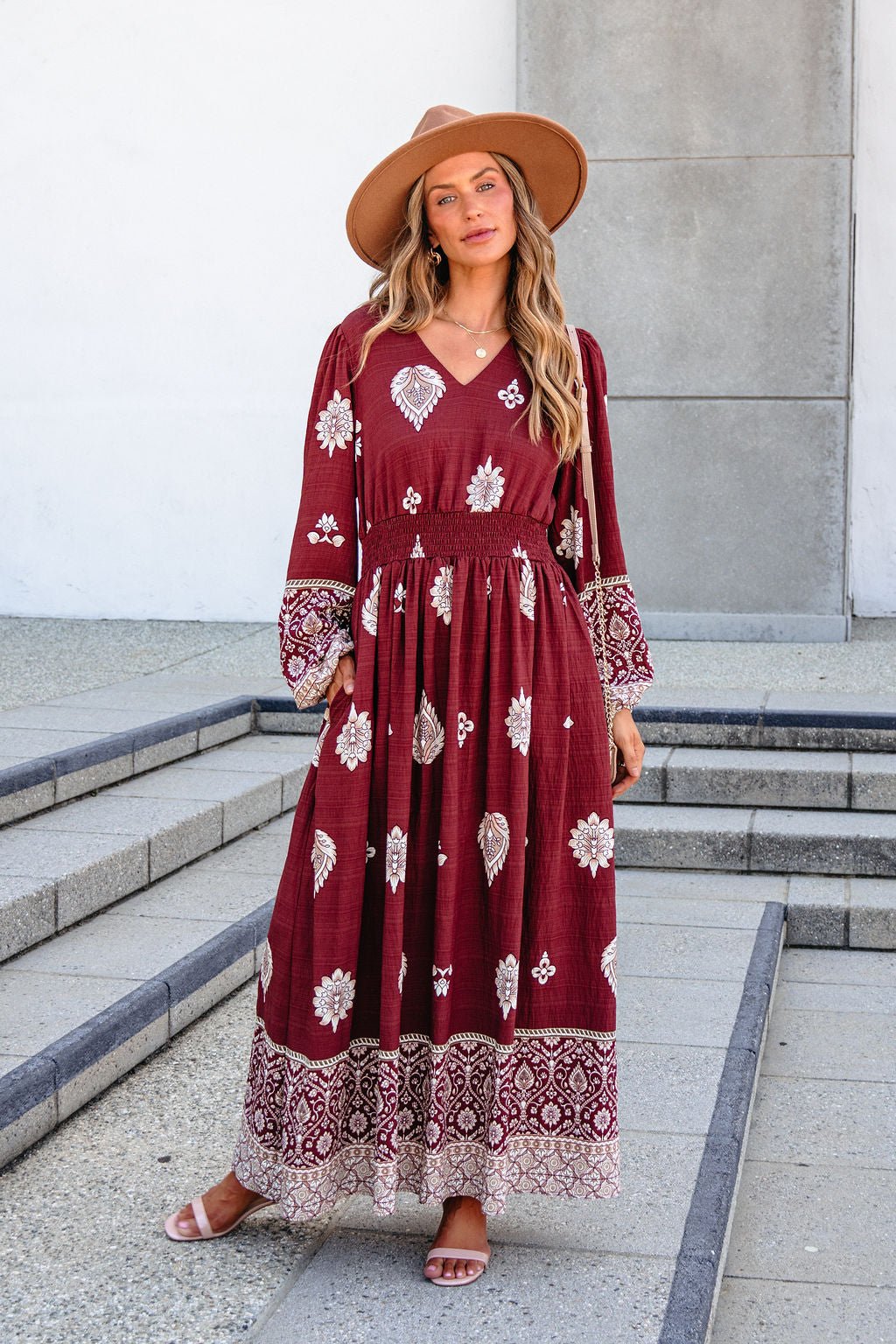 Woman in a SHE AND SKY ST2144 brown printed maxi dress, tan hat, and sandals stands on outdoor steps by a gray and white building.