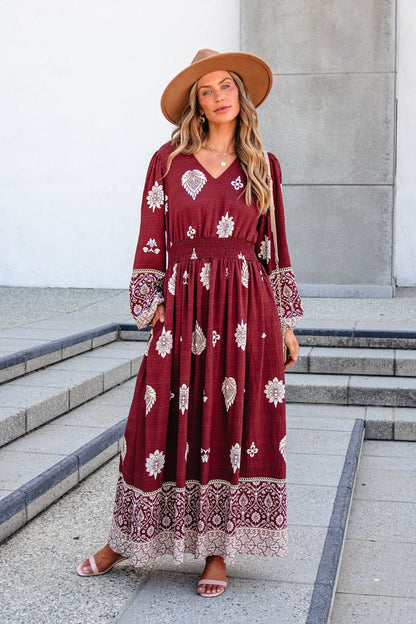 Woman in a SHE AND SKY ST2144 brown printed maxi dress, tan hat, and sandals stands on outdoor steps by a gray and white building.