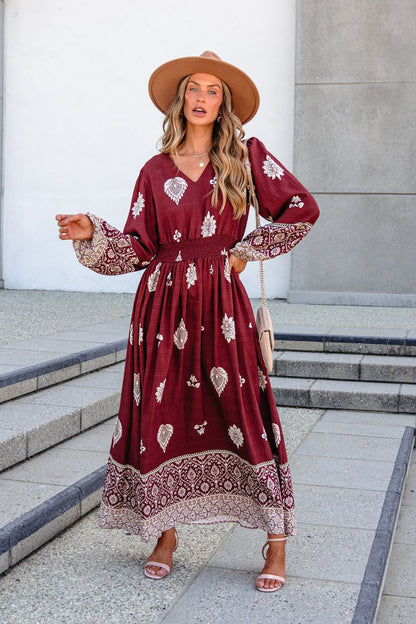 Woman in SHE AND SKY ST2144 brown printed long sleeve maxi dress poses outdoors, holding a beige purse and wearing a tan hat.