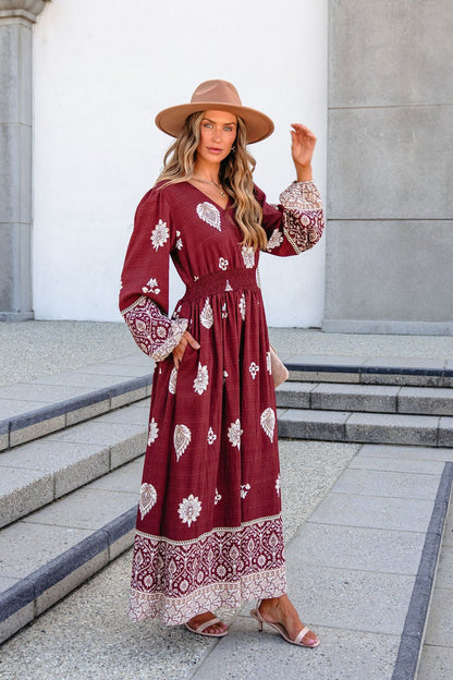 Woman in SHE AND SKY ST2144 brown printed long sleeve maxi dress, tan hat, and sandals poses on outdoor steps with one hand raised.