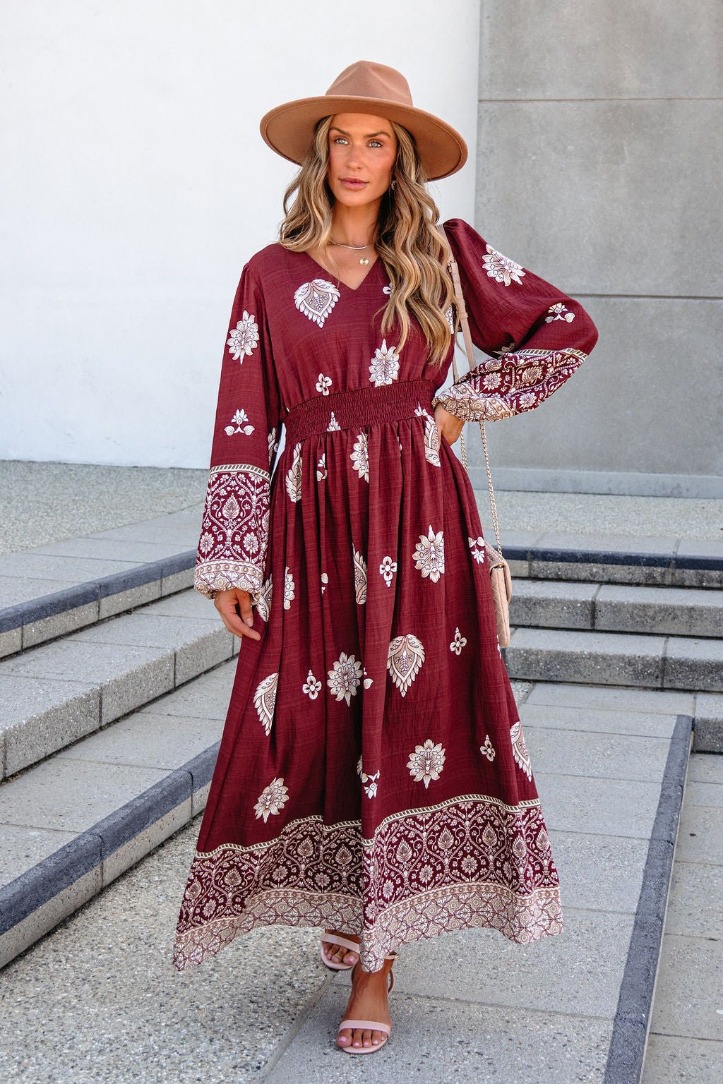 Woman in a SHE AND SKY ST2144 brown printed maxi dress, tan hat, and sandals stands on steps with one hand on her hip.