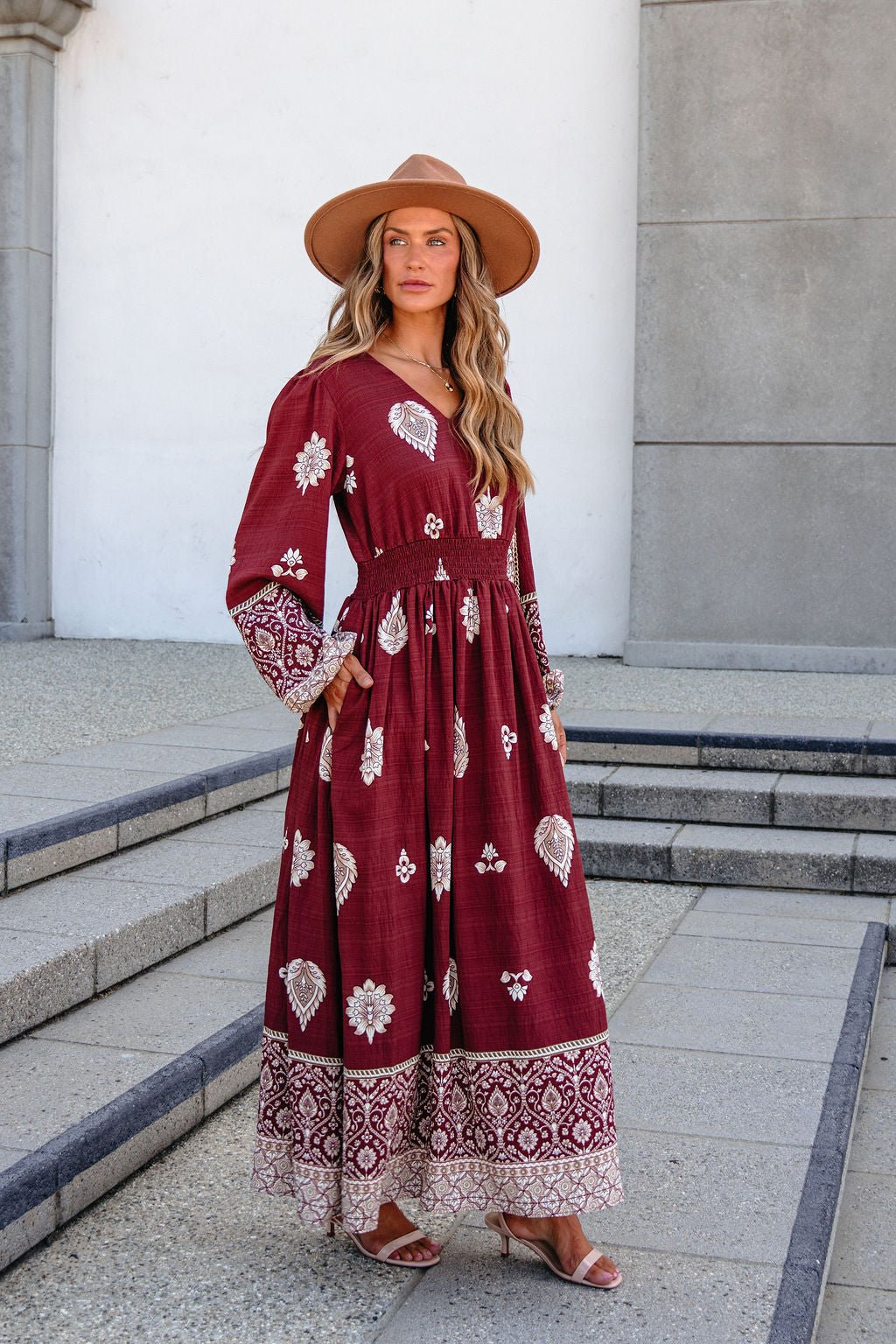 Woman in SHE AND SKY ST2144 brown printed maxi dress, tan hat, and sandals stands on steps outside a gray building.