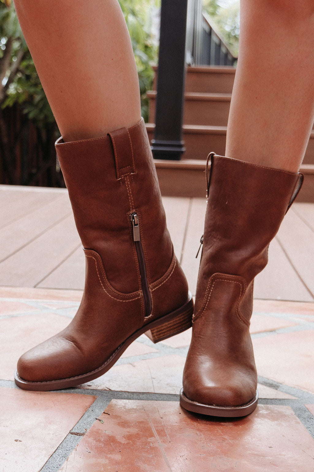 Someone in MIA Miley Mid Calf Boots - Tan stands on tiled flooring near wooden outdoor steps.