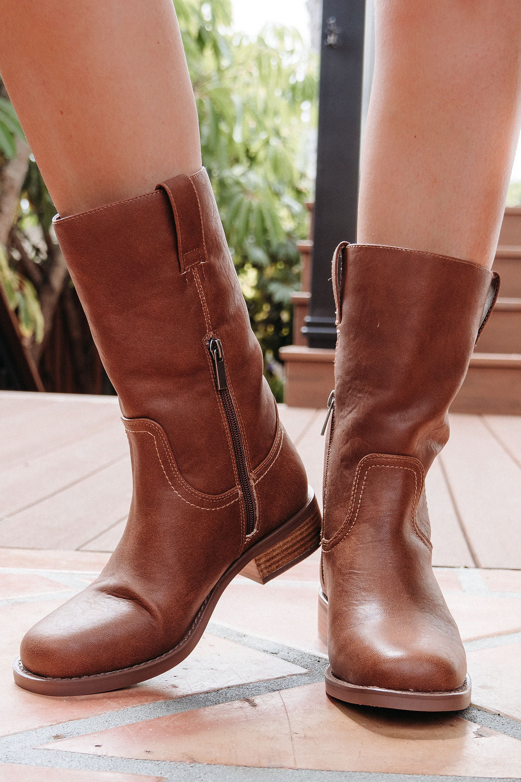 A person wears MIA Miley Mid Calf Boots in tan with side zippers while standing on a wooden deck.