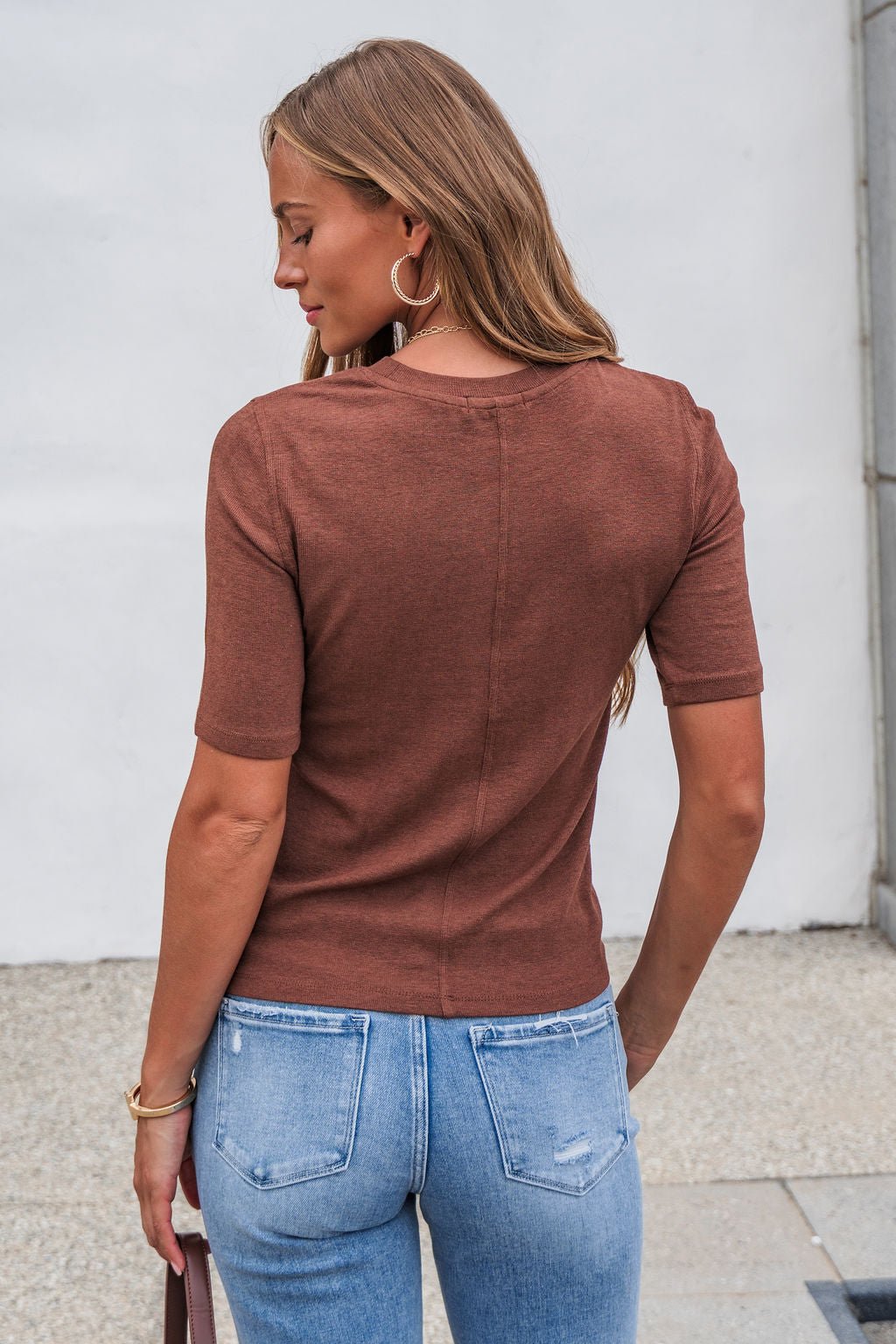 Woman with long brown hair wears a rust short sleeve crew neck sweater and light blue jeans, standing outdoors facing away.