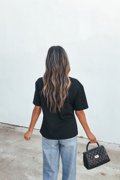 Woman with long wavy hair in the Short Sleeve Metal Trim Top - Black and jeans holds a black handbag, standing by a light wall.