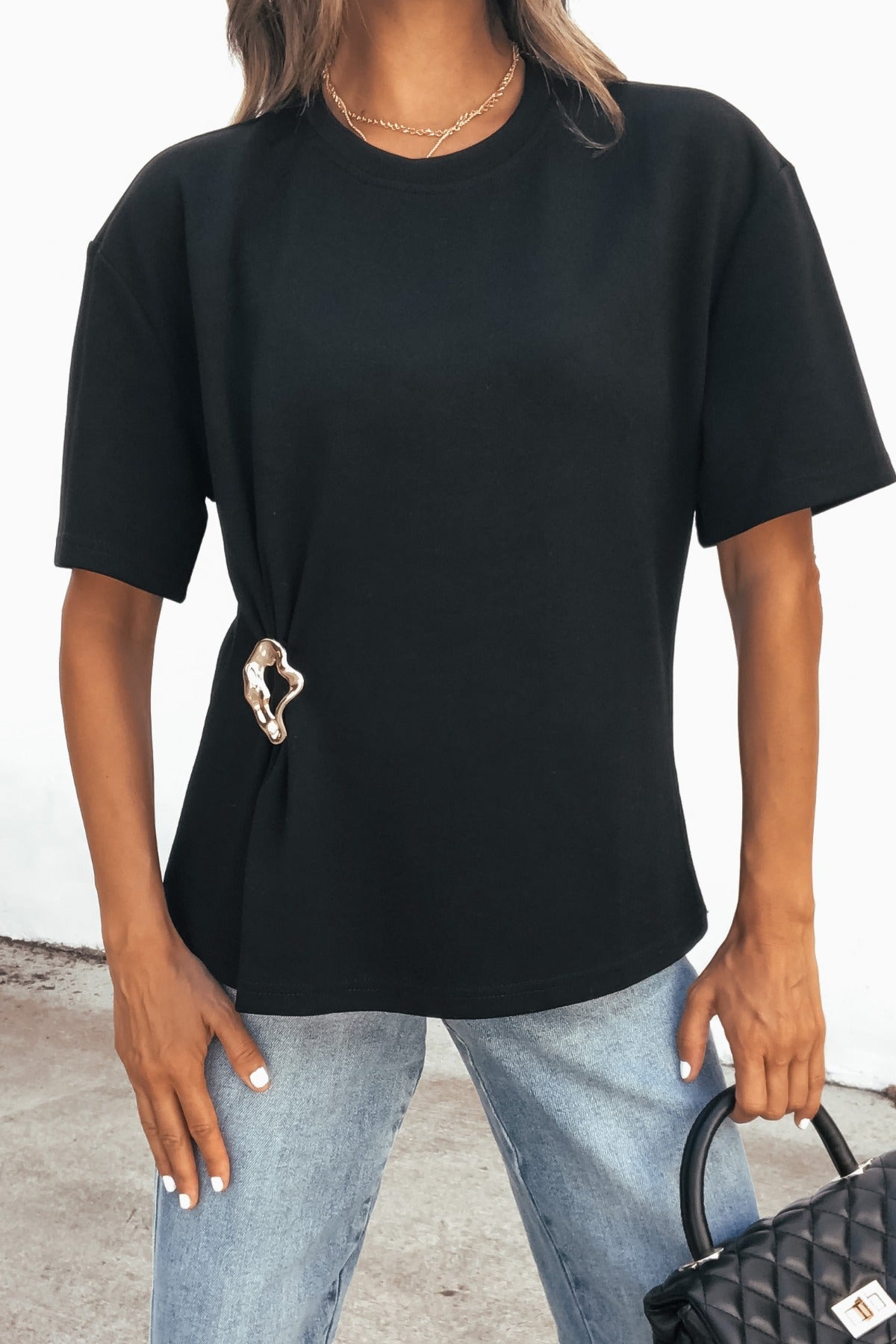 Woman wears a Short Sleeve Metal Trim Top - Black, light blue jeans, and carries a black quilted handbag.