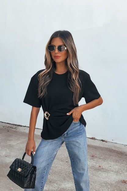 Woman in Short Sleeve Metal Trim Top - Black, blue jeans, black purse, and sunglasses stands by a light wall.