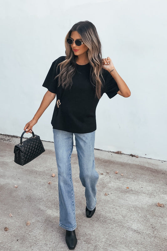 Woman in sunglasses, Short Sleeve Metal Trim Top - Black, light blue jeans, black boots, quilted bag, strolling on concrete.
