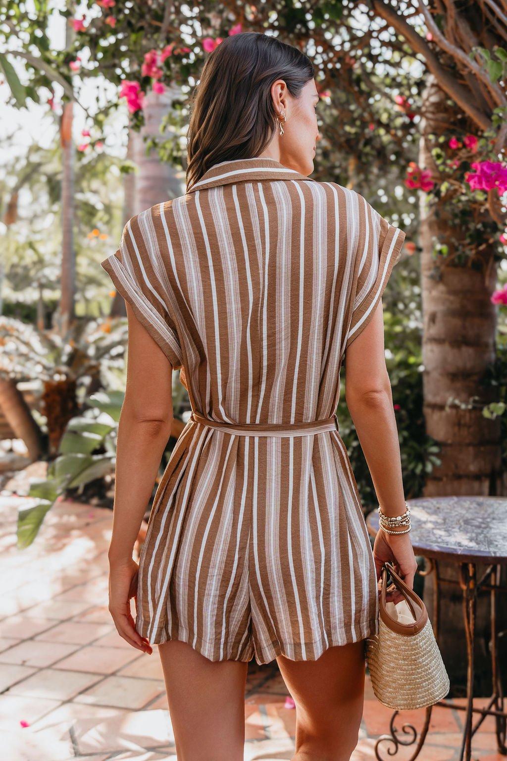 A woman in the Sienna Tan Stripe Button Down Romper stands outdoors by pink flowers, holding a small woven handbag.