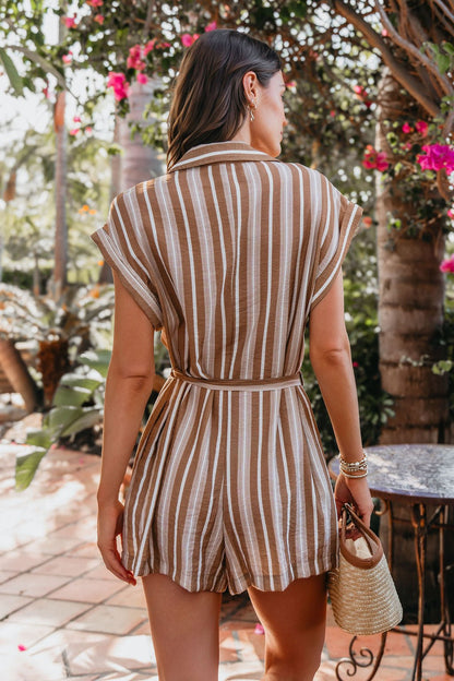 A woman in the Sienna Tan Stripe Button Down Romper stands outdoors by pink flowers, holding a small woven handbag.