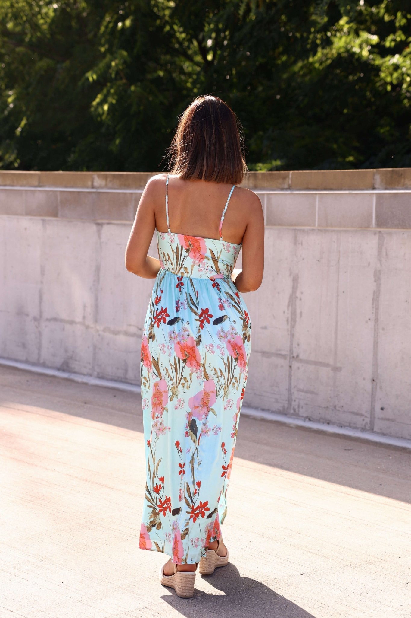 A woman in the Silky Blue Multi Floral Print Maxi Dress and sandals walks outdoors on a sunny day, facing away from the camera.
