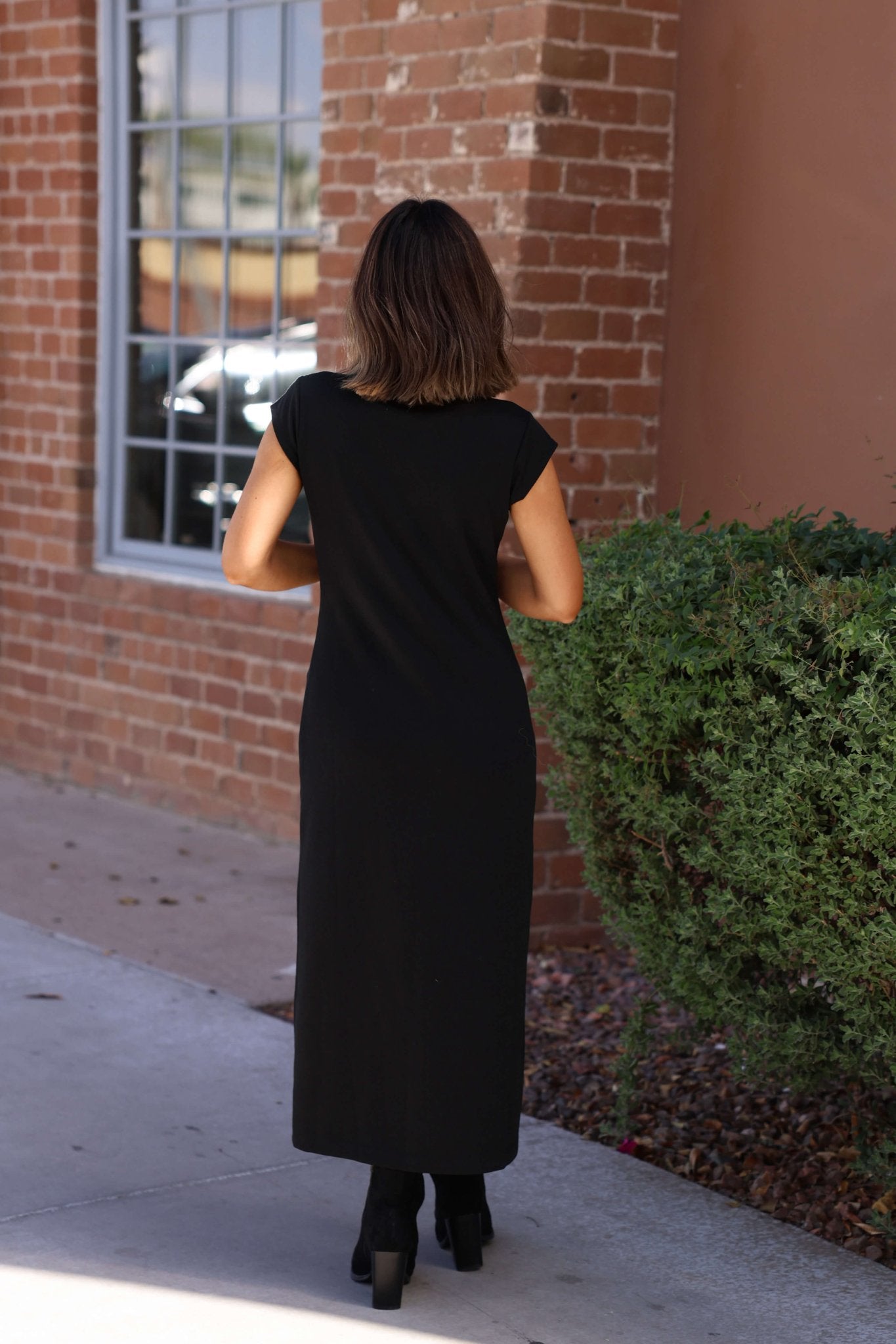 A woman in the Simple Timeless Black Midi Dress stands outside near a brick wall and green shrub, facing away.
